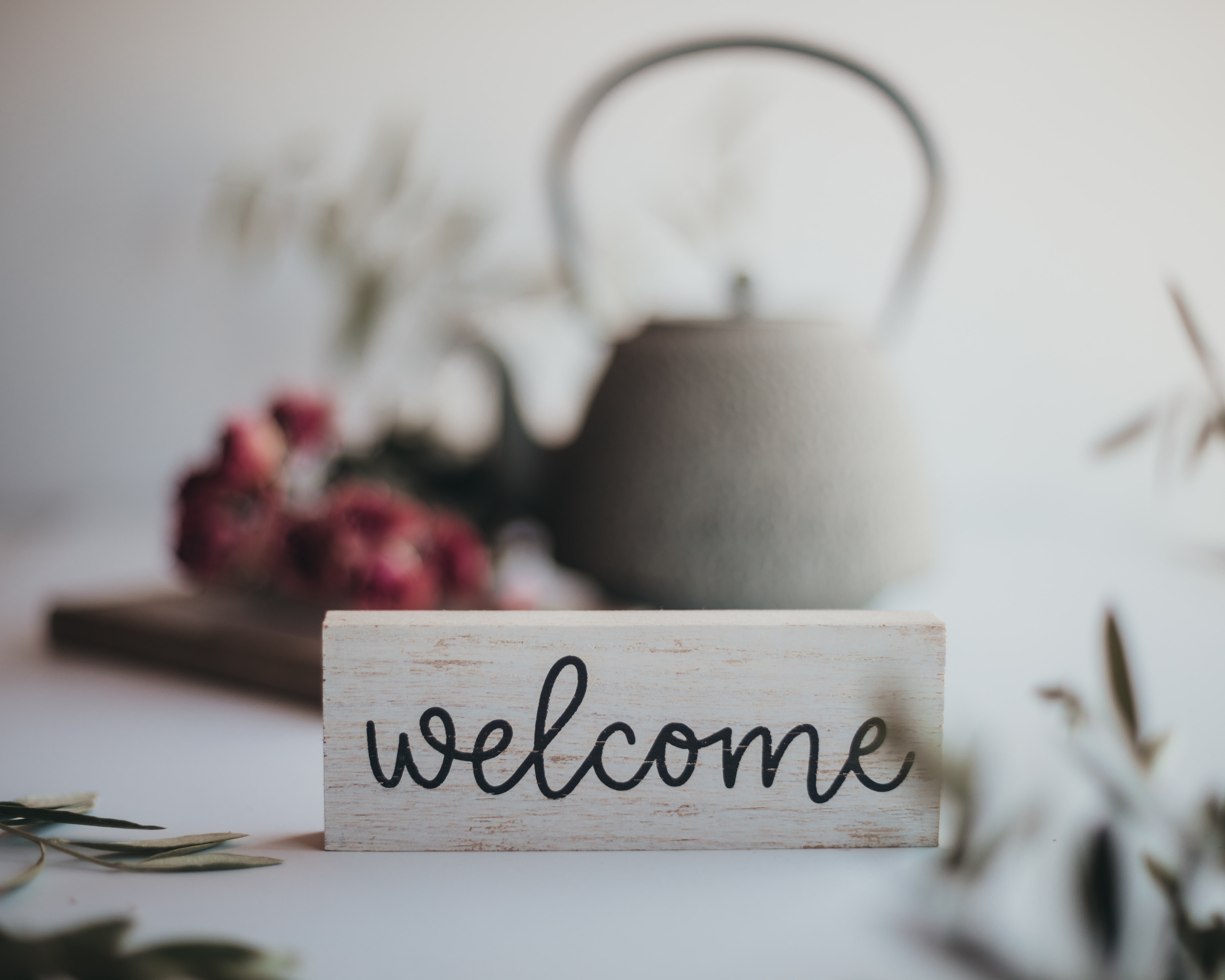 Black cursive 'welcome' written on a piece of white wood in the foreground, with an old fashioned kettle and red flowers blurred in the background.
